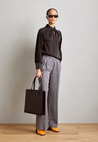 Schwarze Satinbluse mit SchlüssellochDETAIL, graue weiter geschnittene Hose, orange blockabsatz Schuhe und eine schwarze Tragetasche mit Logo.