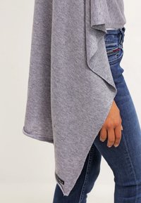 Gros plan d'une personne portant un jean bleu et un châle gris tricoté drapé sur son bras devant un fond uni.