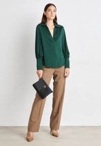 Blusa verde con cuello de tapeta y mangas abullonadas; combinada con pantalones marrones de pierna recta. Sostiene un bolso de mano negro con patrón de espiguilla.