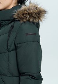 Grønne pufferjakke med en faux fur-hætte, quiltet tekstur, justerbare snore og et sort logo på ærmet.