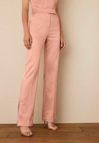 Pantalons en tissu doux rose pâle, coupe ajustée avec une jambe droite et de subtiles fentes latérales à l'ourlet. Associés à des talons dorés à lanières.