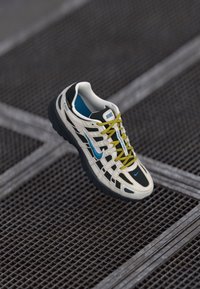 Baltās un melnās Nike skriešanas kurpes ar zilu Swoosh un dzelten�ām šņorēm, izstādītas uz tumšas metāla režģa virsmas.