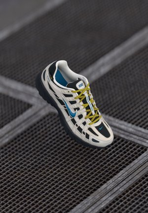 Zapatilla de running Nike en blanco y negro con swoosh azul y cordones amarillos, exhibida sobre una superficie de rejilla de metal oscuro.