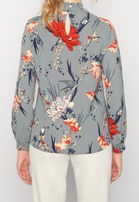 Blommig blus i ljusgrått tyg med röda, rosa och blå blommönster. Har långa ärmar, rundad krage och en nyckelhålsdetalj på baksidan.