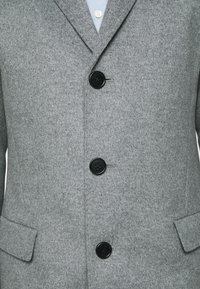 Cappotto grigio in lana con tre bottoni neri allacciati, che mostra i risvolti, una camicia azzurra chiara sotto e tasche a pattina su entrambi i lati.