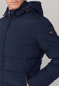 Chaqueta acolchada azul marino con cierre de cremallera, capucha con cordones ajustables y bolsillos laterales. Presenta un diseño acolchado y una parche de marca en la manga.