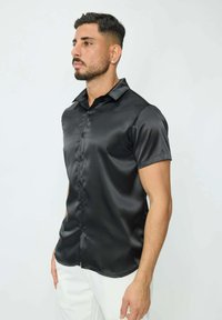 Chemise noire en satin à manches courtes avec un devant à boutons, texture lisse et col classique, portée avec un pantalon blanc.