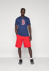 T-shirt della Marina con logo rosso di Boston, abbinato a pantaloni corti rossi. Made in cotone, presenta maniche corte e collo rotondo. Sneakers nere completano l'outfit.