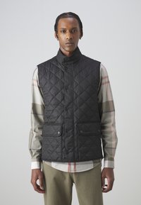 Colete preto acolchoado sem mangas com gola alta, apresentando grandes bolsos frontais e fechos de botão de pressão, usado sobre uma camisa de manga longa xadrez.