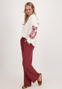 Witte textuurblouse met een rode geborduurde bloemenpatroon op de mouwen, gecombineerd met bordeauxwijde broek en bruine sandalen met bandjes.