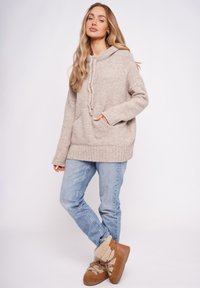 Beżowy sweter z dzianiny z kapturem, kieszenią z przodu i troczkami, zestawiony z jasnoniebieskimi dżinsami i brązowymi botkami wykończonymi futerkiem.