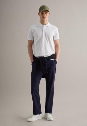 Un giovane uomo indossa una polo bianca, pantaloni blu navy, scarpe da ginnastica bianche, un maglione nero legato intorno alla vita e un cappello verde oliva su uno sfondo neutro.