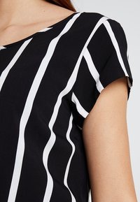 Černobílý pruhovaný top s krátkými rukávy, kulatým výstřihem a hladkou texturou látky. Proužky jsou zvlněné a různé šířky.