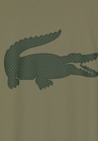 Graphic di un coccodrillo verde su sfondo khaki, con squame texture e una forma slanciata ed elegante.