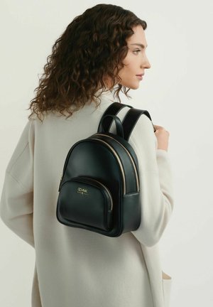 Kobieta o kręconych włosach, ubrana w kremowy płaszcz, niesie na ramieniu mały czarny skórzany plecak ze złotymi zamkami błyskawicznymi i logo.