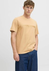 Bež pamučna majica kratkih rukava s blagim reljefnim logotipom "JACK & JONES PREMIUM" na prsima, u kombinaciji s tamnoplavim trapericama.