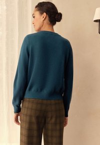 Pleteni pulover v turkizni barvi z dolgimi rokavi, okroglim izrezom in rebrastim robom, kombiniran s hlačami rjave kariraste tkanine. Mehka tekstura, sproščen kroj.