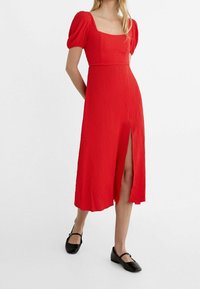 Punainen teksturoitu midi-mekko, jossa on puffihihat, neliönmuotoinen pääntie ja etulahje, yhdistettynä mustiin tasapohjaisiin kenkiin.