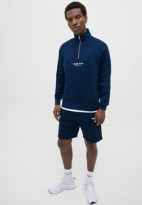Sudadera con cremallera color azul marino con logo, combinada con pantalones cortos a juego. La tela es suave y el diseño presenta un corte relajado y acentos en blanco.