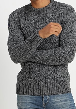 Homme portant un pull gris en maille torsadée avec poignets et ourlet côtelés, associé à un jean bleu en denim, bras croisés.