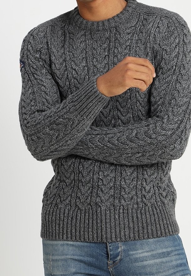 Homme portant un pull gris en maille torsadée avec poignets et ourlet côtelés, associé à un jean bleu en denim, bras croisés.