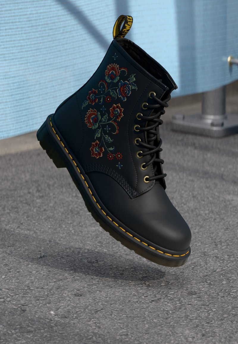 Bottes en cuir noir à lacets avec coutures jaunes et broderie florale rouge, flottant au-dessus de l'asphalte près d'une barrière bleu clair.