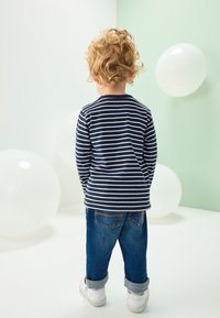 Langærmet skjorte med blå og hvide striber kombineret med opkrøllede denimjeans. Barnet vender ryggen til, med hvide balloner i baggrunden.