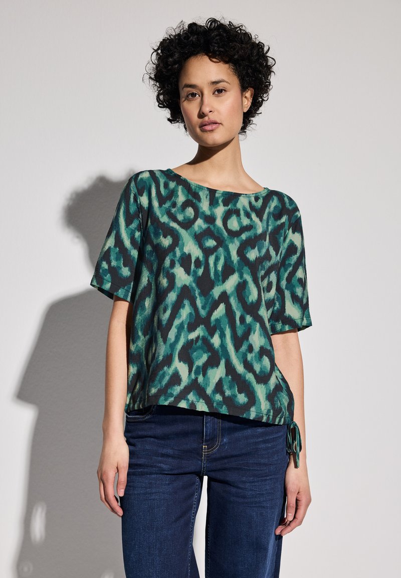 Maglietta ampia in stampa animalier verde e nera, con maniche corte, scollo a barca e un dettaglio con fiocco laterale. Tessuto morbido.