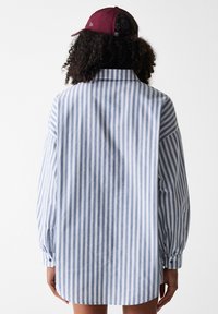 Light blue and white striped oversized shirt with long sleeves, soft cotton material, and a curved hem. Worn with a maroon cap.