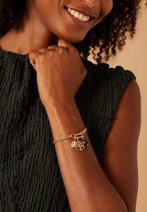 Smiling person with curly hair wears a textured black sleeveless top and a gold chain bracelet with three charms on their wrist.