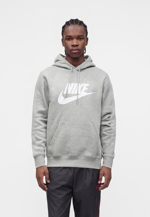 Młody mężczyzna z warkoczami, ubrany w szarą bluzę z kapturem Nike i czarne spodnie Nike, stojący na jednolitym białym tle.