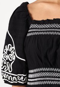 Svart bluse med puffede ermer og hvit broderi. Fremragende detaljer inkluderer intrikat søm over bryststykket og ermelige aksenter.
