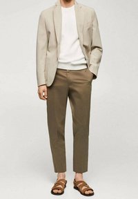 Blazer beige su una camicia bianca a costine, abbinato a pantaloni khaki a sigaretta e sandali marroni con cinturini. Design semplice e pulito.