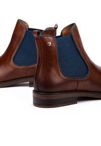 Brown leather Chelsea boots with dark blue elastic side panels and low stacked heels on a white background.