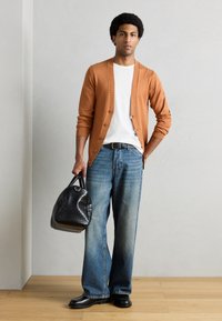 Πλεκτή πορτοκαλί ζακέτα, λευκό t-shirt, χαλαρά μπλε τζιν και μαύρα δερμάτινα παπούτσια. Κρατάει μια μαύρη τσάντα με υφή. Ξύλινο πάτωμα και ουδέτερος τοίχος.