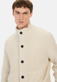 Beige strikket cardigan med hævet tekstur, høj krave og sorte knapper. Har to forlommer for ekstra detaljer.