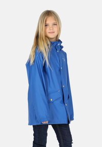 Jeune fille aux longs cheveux blonds portant une veste imperméable à capuche bleue et un jean foncé, debout devant un fond clair uni.