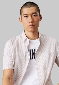 Camicia a maniche corte color beige chiaro a righe con bottoni e taschino, indossata sopra una t-shirt bianca con lettering nero.