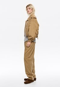 Una modella è di profilo, indossando un giubbotto oversize beige, un maglione grigio e pantaloni larghi, con scarpe marroni, su uno sfondo bianco.