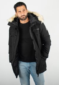 Parka noire avec capuche en fausse fourrure, fermetures éclair et boutons, plusieurs poches et une texture lisse. Présente un logo sur la poitrine.