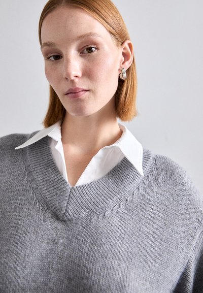 Pulover tricotat de culoare gri deschis, cu un design în formă de V, având modele texturate și manșete cu nervuri, purtat peste o cămașă albă cu guler.