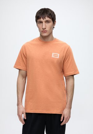 Jeune homme aux cheveux courts et foncés portant un T-shirt orange uni avec un petit logo rectangulaire blanc et orange sur la poitrine, debout devant un fond blanc.