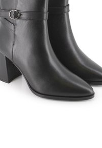 Botas de tobillo de cuero negro con punta afilada, tacón cuadrado y una correa decorativa que presenta una hebilla metálica circular. Textura suave en toda la superficie.
