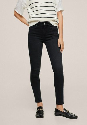 Frau trägt schwarze Skinny-Jeans, weißen kurzärmeligen gestreiften Pullover und schwarze Leder-Loafers, steht vor neutralem Hintergrund.