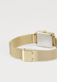 Bracelet de montre en métal doré en maille avec fermoir et boîtier de montre carré, vu de l'arrière sur un fond blanc.