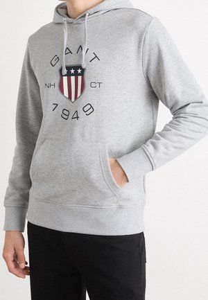 Person trägt einen hellgrauen Hoodie mit einem roten, weißen und blauen Schild-Logo und dem Schriftzug GANT 1949 auf der Brust.