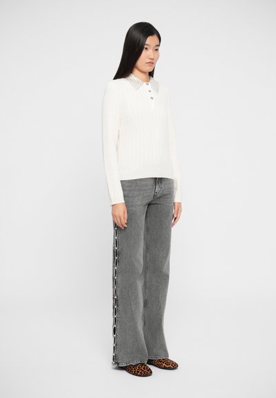 Ung kvinde iført en hvid strikket sweater med dekoreret krave, grå brede jeans med kædedetaljer på siderne og sko med leopardprint.