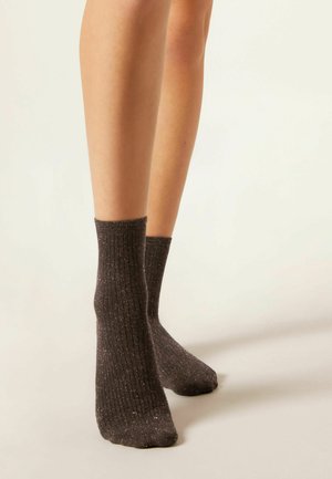 Des jambes portant des chaussettes côtelées marron foncé avec de petites paillettes argentées, se tenant sur un fond clair et uni.