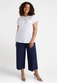 Top blanco de manga corta con un lazo decorativo plateado en el escote, combinado con pantalones de pierna ancha azul marino y tacones plateados con tiras.