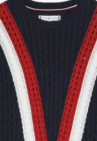 Granatowy sweter z plecionki w sploty, z czerwono-białym wzorem w kształcie litery V i etykietą Tommy Hilfiger przy kołnierzu.